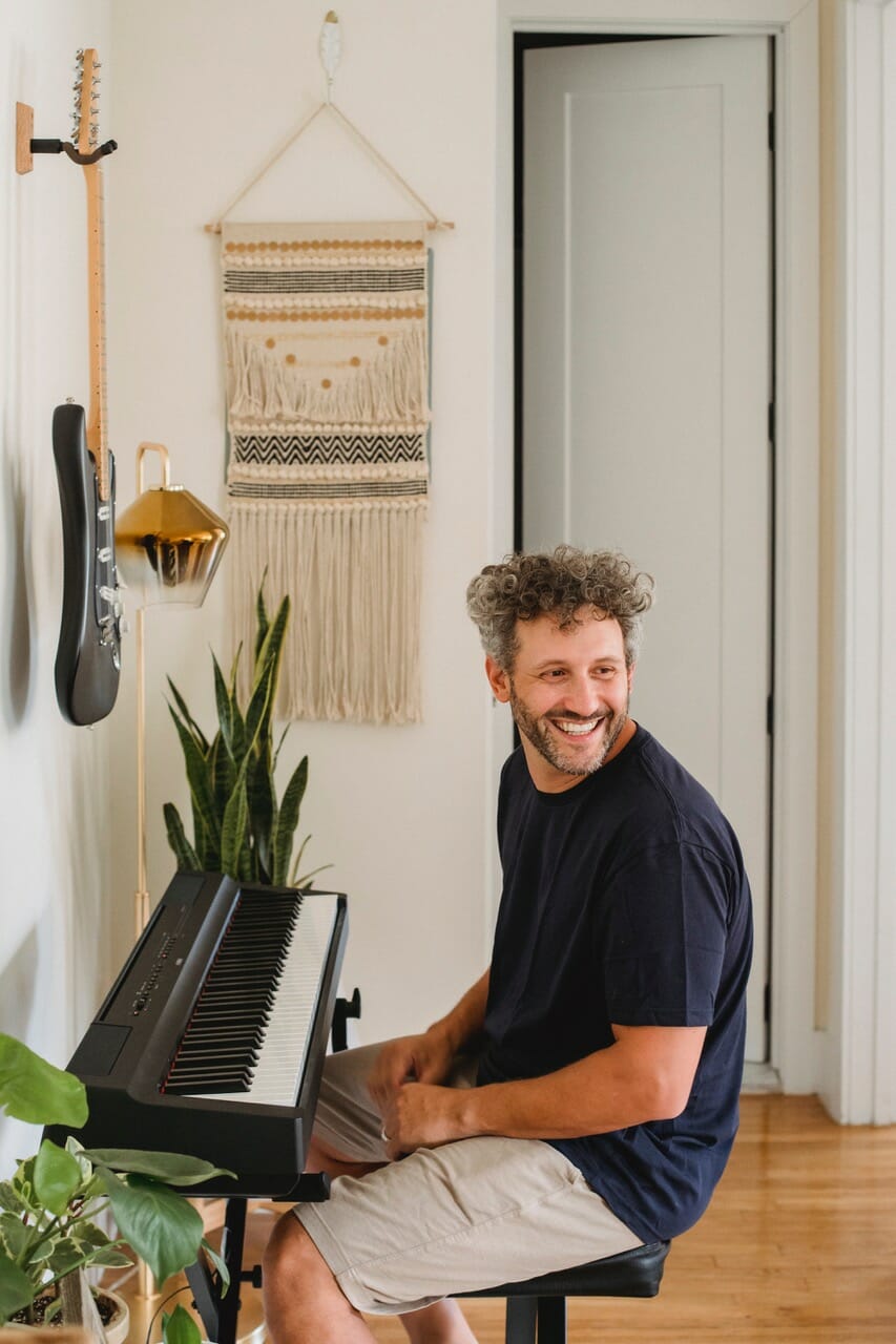 Adult Man Playing music at a Piano with Electric Guitar on the Wall
